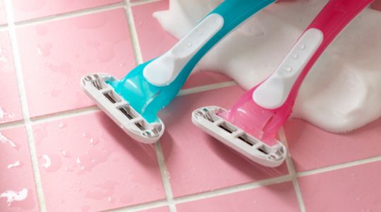 Two disposable razors, one blue and one pink, lying on a wet pink tiled surface with shaving foam around them.